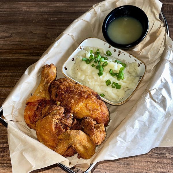 Fried chicken, mashed potatoes with chives, and a side of gravy in a basket on a wooden table.