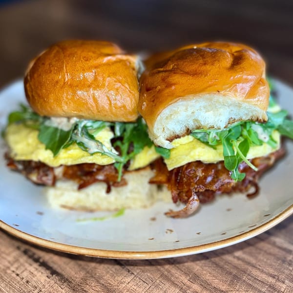 Two sliders with scrambled eggs, arugula, and pulled pork on brioche buns, on a white plate.