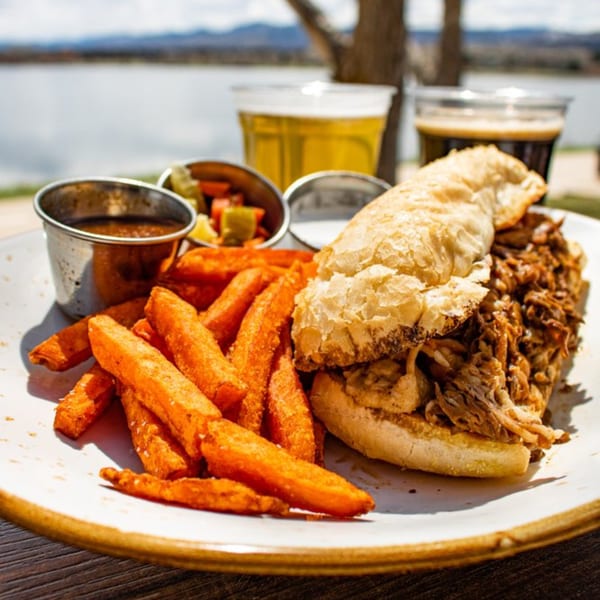Pulled pork sandwich with sweet potato fries, sauces, and two beers by a lake.
