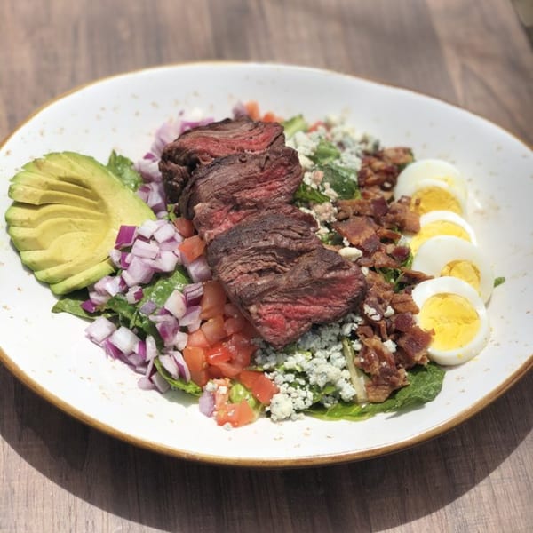 A steak salad with avocado, red onion, tomatoes, blue cheese, bacon, and hard-boiled eggs.