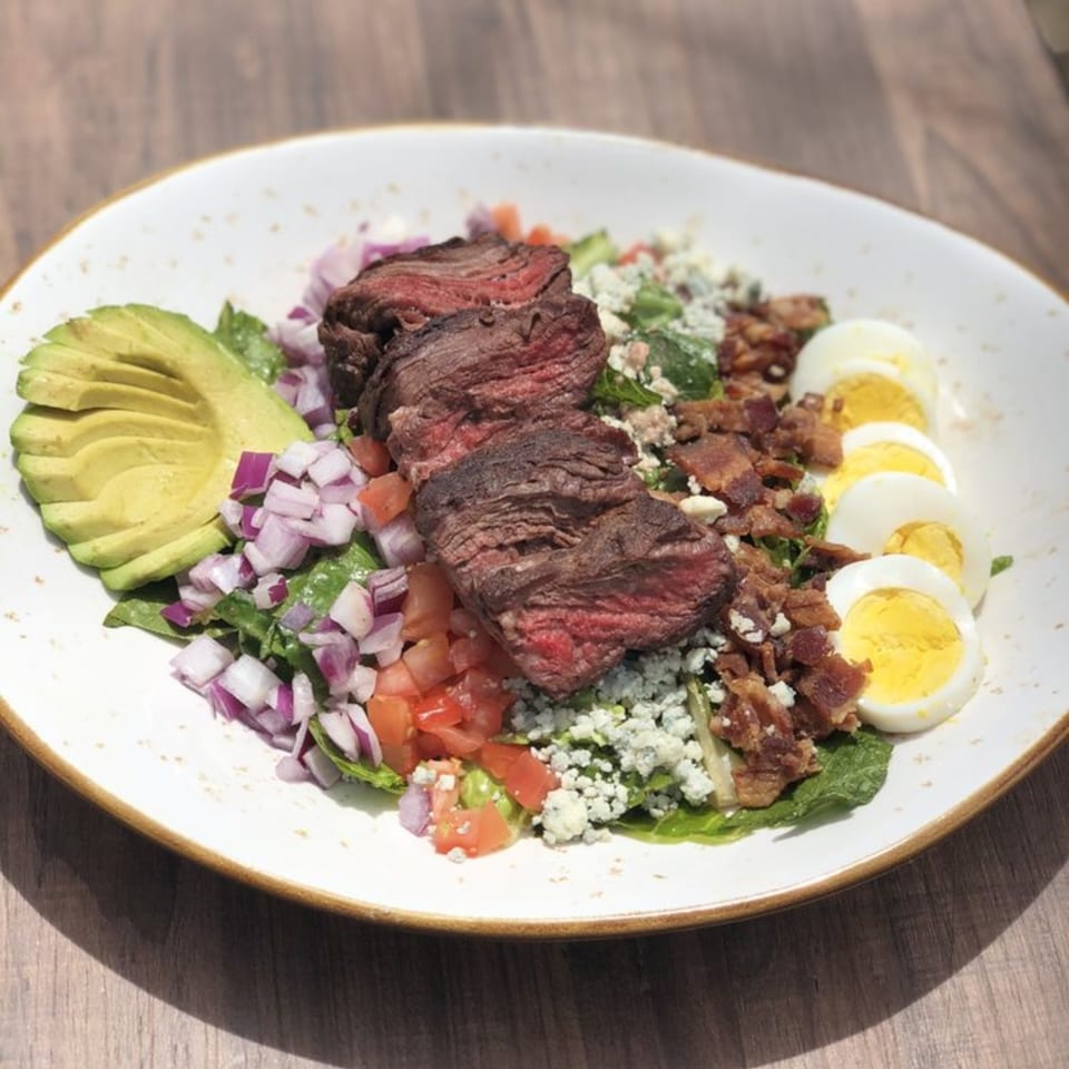 A steak salad with avocado, red onion, tomatoes, blue cheese, bacon, and hard-boiled eggs.